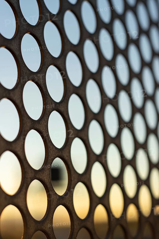 Vertical view of the sunset through a perforated steel sheet with holes in the shape of circles. Selective focus, AI generated