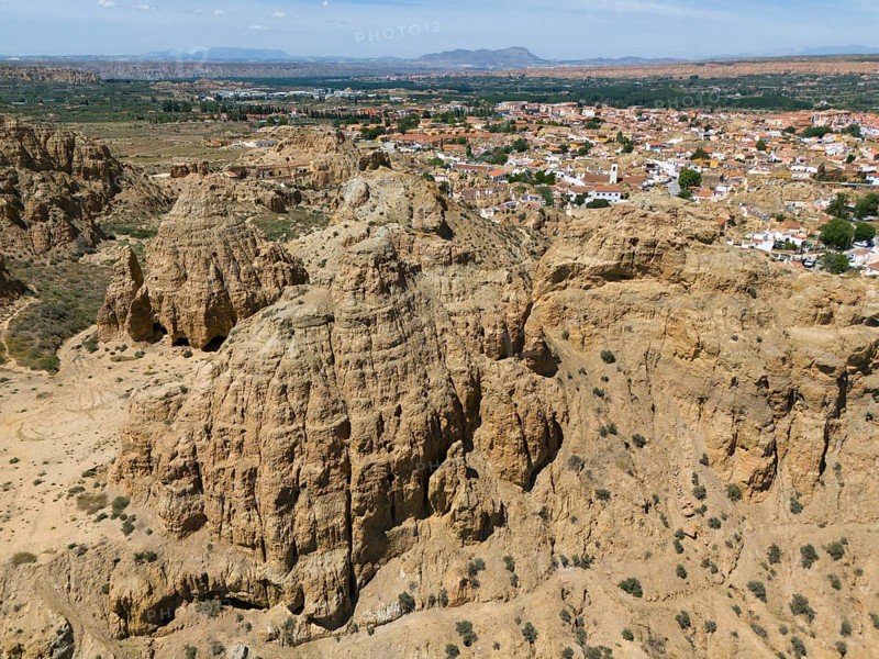 Imposing rock formations in front of a wide, bare landscape with a small village in the distance, aerial view, cave dwellings, cave district, Troglodytos, Guadix, Granada, Andalusia, Spain, Europe