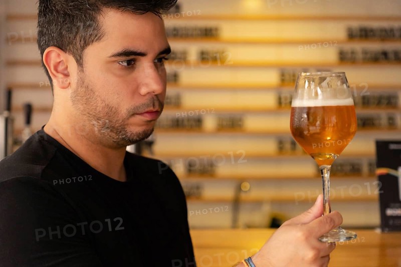A young Latino man enjoys a craft beer in a pub. He carefully observes the color and hops in the lager he is tasting, showing his appreciation for good brewing and craft beer culture