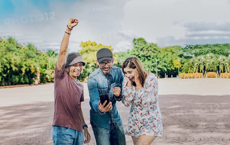 Three cheerful friends looking at cell phone making gesture winner outdoors. Excited teenage friends looking at cell phone screen on the street