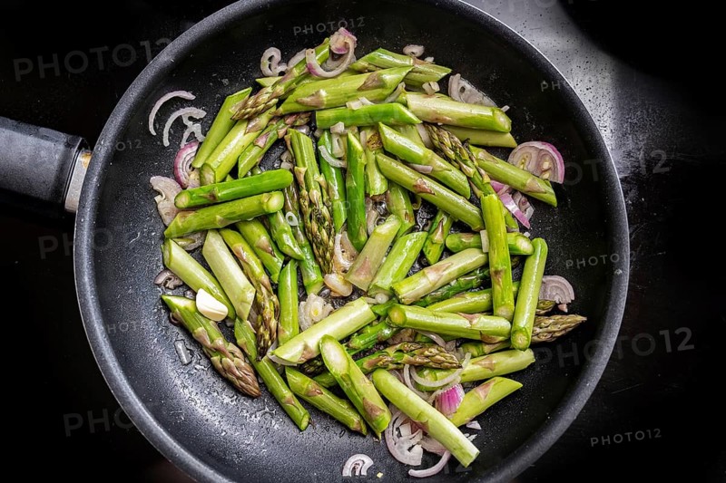 Green asparagus and red onion in a frying pan on the black stove top, cooking a healthy vegetarian meal with seasonal vegetables, high angle view from above, selected focus