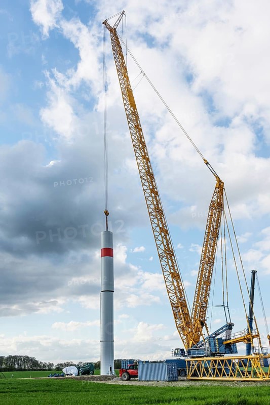 Giant crane is moving a tower part from steel of a wind turbine onto the first tube, heavy industry construction site for a power plant for renewable energy, dark clouds, blue sky, copy space, selected focus