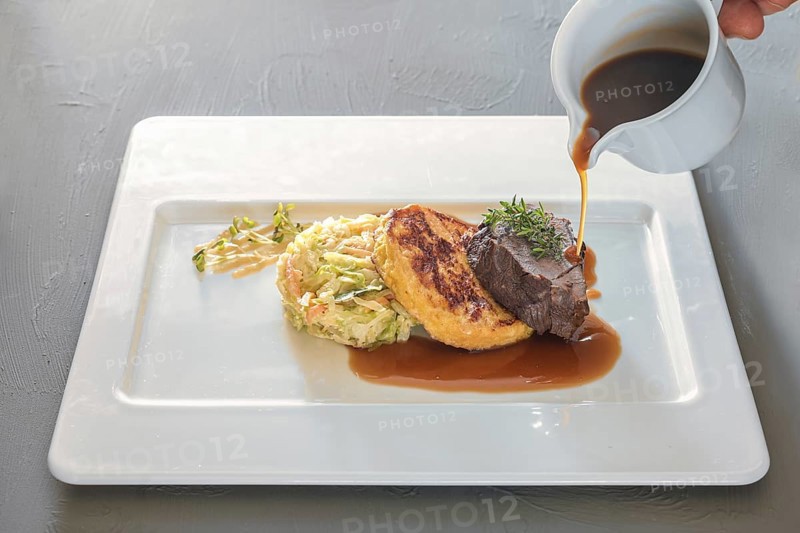 Pouring sauce on a dish from roast venison, flat bread dumpling and round shaped cabbage vegetable on a white plate, preparation for delicious gourmet meal, copy space, selected focus