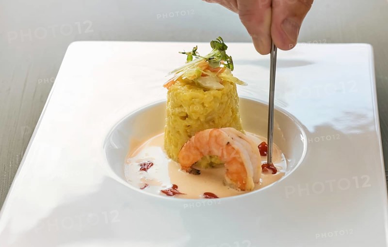 Hand with tweezers garnishing a gourmet dish from saffron risotto with herb topping, a large prawn and crustacean sauce in a modern white plate, copy space, selected focus, narrow depth of field