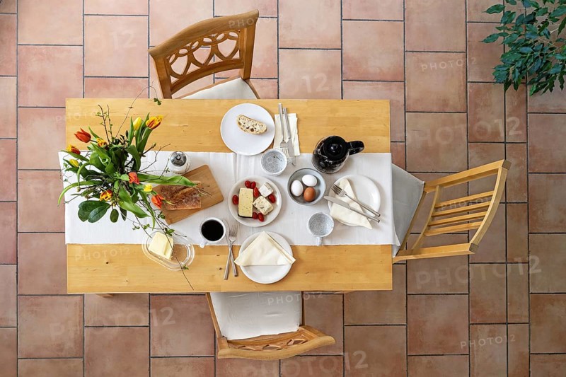 Breakfast on a wooden table with bread, cheese, eggs and coffee and a bouquet of tulips as decoration, three vintage chairs on a tiled floor, high angle view from above, selected focus