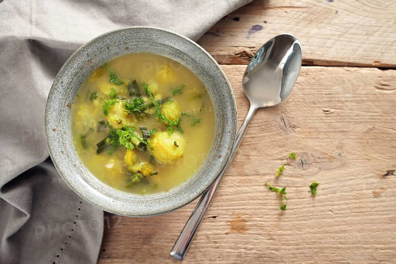 Vegetable soup with brussels sprout, leek and potato in a bowl, spoon and napkin on a rustic wooden table, high angle view from above, copy space, selected focus