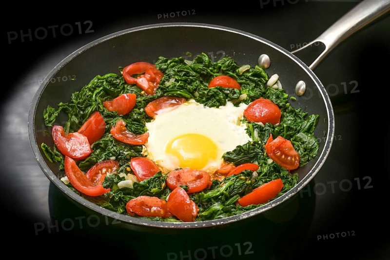 Fried egg with spinach and tomatoes in a frying pan on a black cooker top, preparing a healthy meal for breakfast or lunch, copy space, selected focus, narrow depth of field