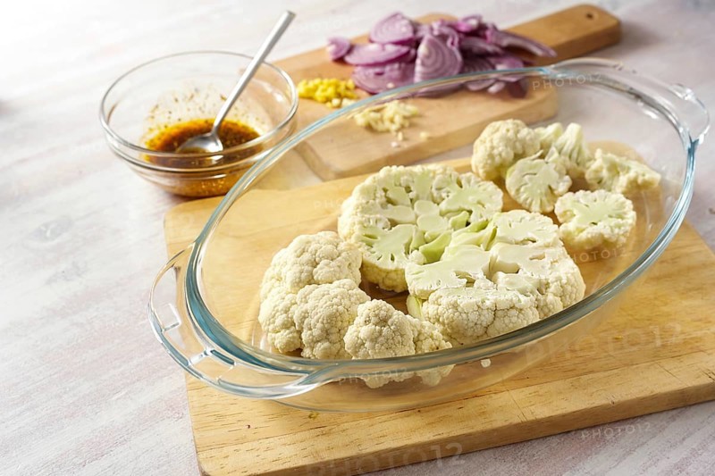 Raw cauliflower slices in a glass casserole, chopped red onion and a spice mix in olive oil, cooking preparation for baked vegetable steaks, copy space, selected focus, narrow depth of field