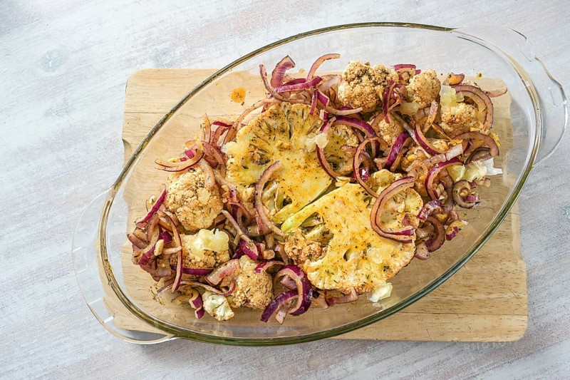 Baked cauliflower steaks with spicy oil and red onions in a glass casserole on a wooden kitchen board on a light table, vegetarian vegetable meal, top view from above, selected focus