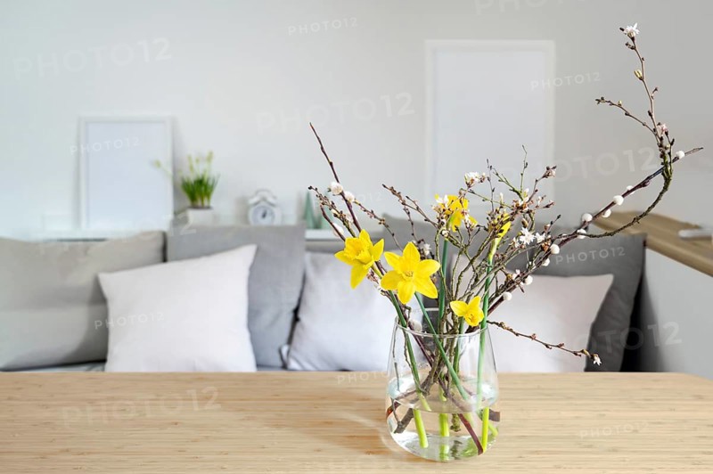 Spring bouquet with daffodils and flowering branches in a glass vase on a table in front of a sofa bench, seasonal home decor, copy space, selected focus, narrow depth of field