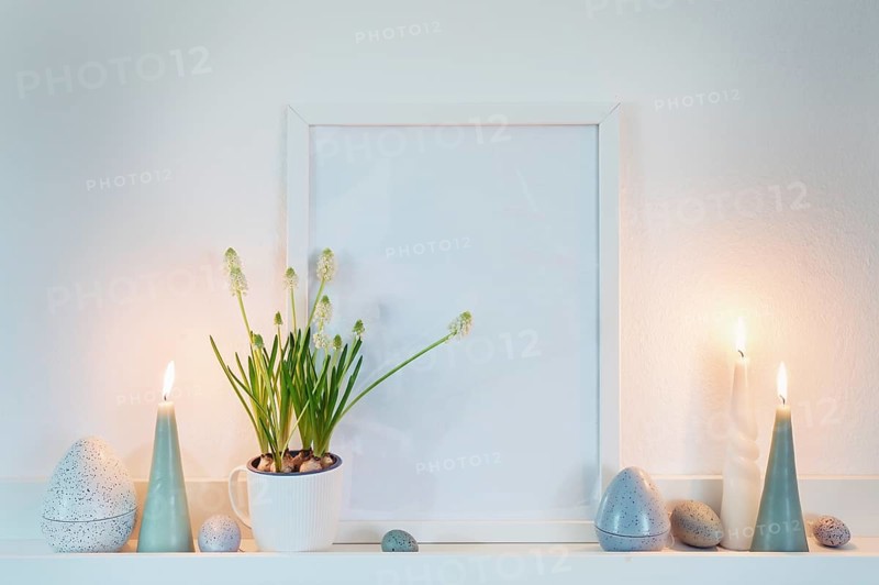 Decoration with an empty picture frame, candles and easter eggs in dusty blue green colors and a potted grape hyacinth plant on a white shelf on the wall, home decor, copy space, selected focus