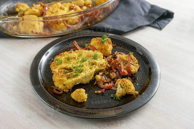 Cauliflower steaks baked with spices, herbs and red onions in a casserole, portion with parsley garnish on a black plate and a gray wooden table, healthy vegetable meal, copy space, selected focus