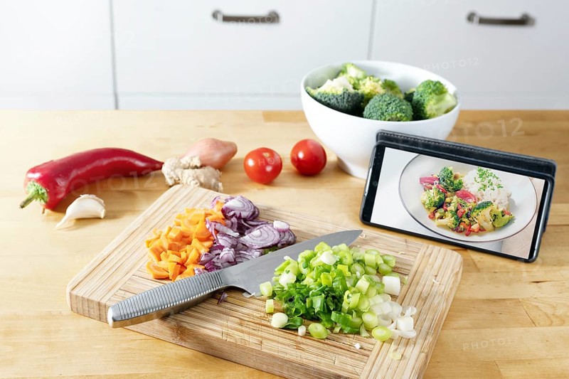 Cut vegetables on a wooden kitchen board, picture of the finished recipe on the smartphone, cooking by using an online app, modern preparation of a healthy meal, selected focus, narrow depth of field