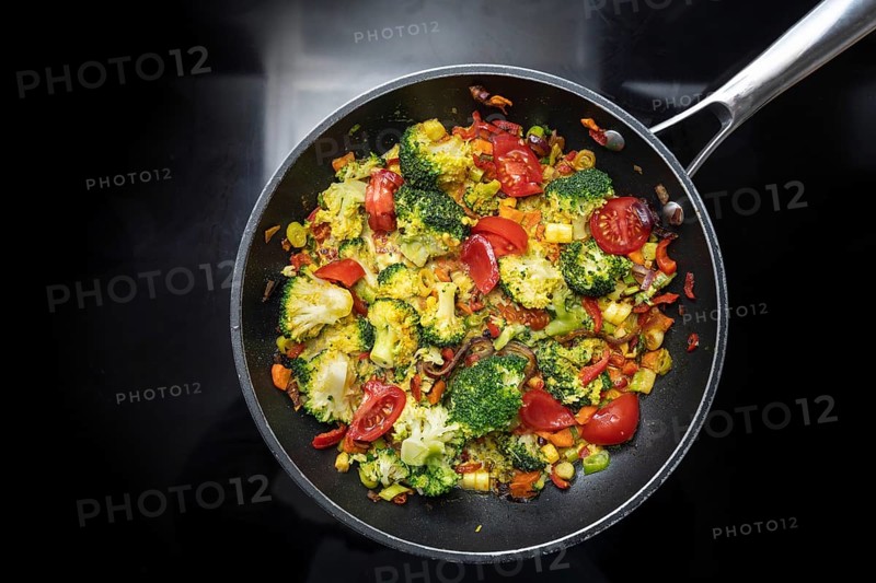 Stir-fried vegetable meal from broccoli, bell pepper, onion and tomato in a frying pan on the black stovetop, vegetarian cooking concept, high angle view from above, copy space, selected focus
