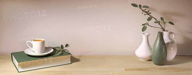 Coffee cup on a gray green book and a group of vases with a sage leaf branch on a wooden table table against a pale wall, panoramic format, home decor, copy space, selected focus
