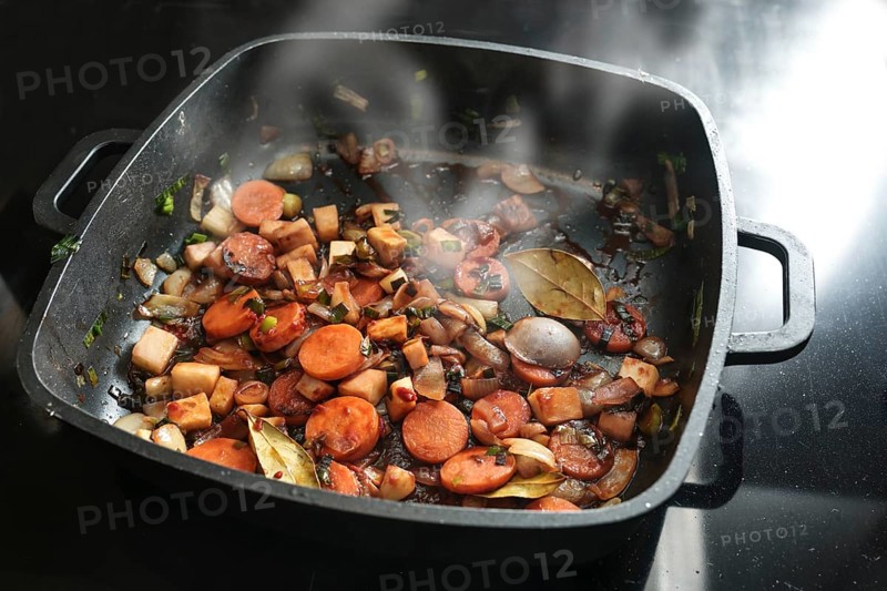 Roasted vegetables for a tasty sauce base in a black cooking pan with carrots, onions, leeks, celery and bay leaves, copy space, selected focus, narrow depth of field