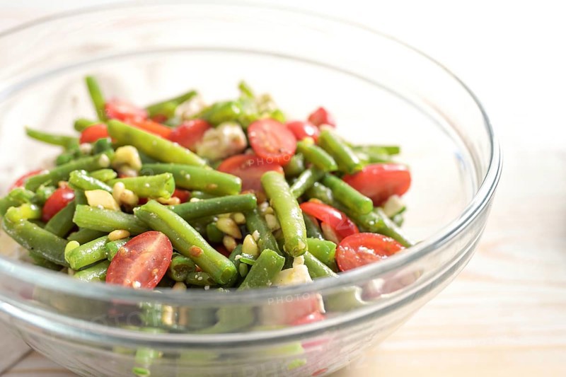 Salad of green beans, tomatoes, pine nuts and feta cheese in a glass bowl on a light wooden table, Mediterranean antipasto, copy space, selected focus, narrow depth of field