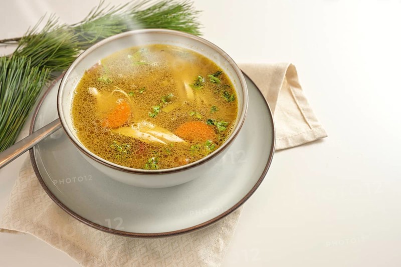 Hot steaming winter soup from chicken, vegetables and parsley in a bowl, home remedy against cold and flu, served on a gay white table with pine branches, copy space, selected focus, narrow depth of field