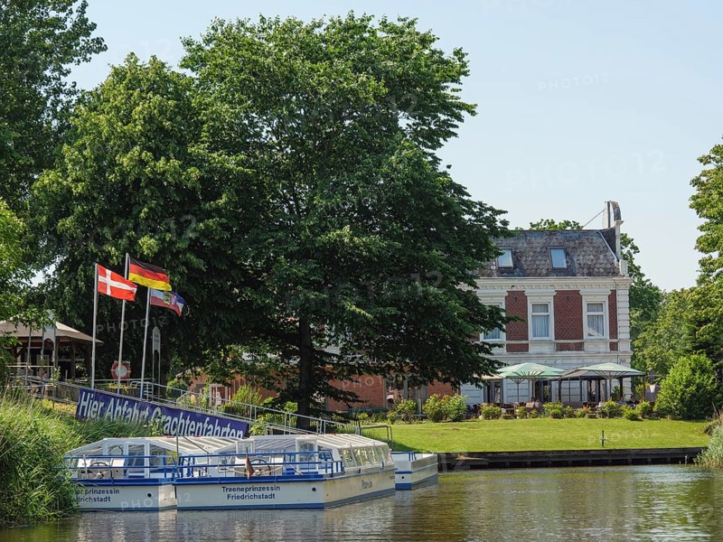 A tourist boat on the canal, surrounded by green trees and a historic villa with flags in the foreground, friedrichstadt, schleswig-holstein, germany