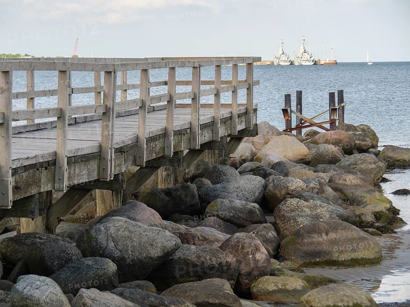 Wooden jetty next to large rocks on the shore with ships in the background, Eckernförde, SChleswig-Holstein, Germany, Europe