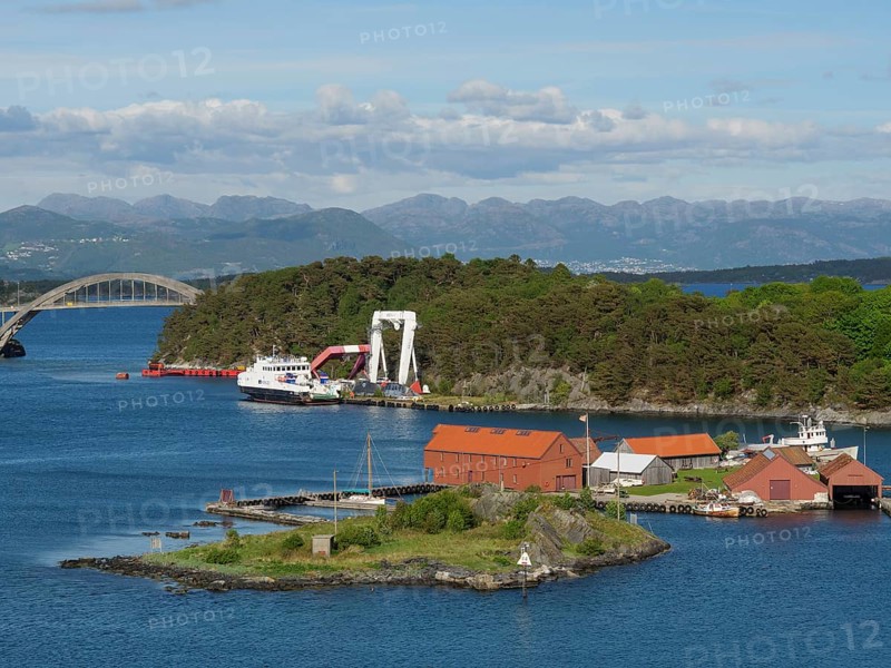 Small island with red buildings and boats, surrounded by the sea. A bridge and mountains are visible in the background, Stavanger, Norway, Europe