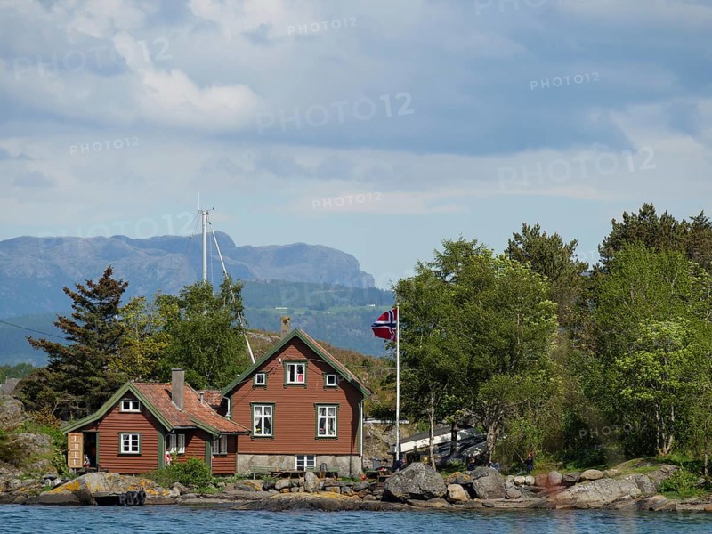 Red wooden house with flag in front of a picturesque landscape with trees, water and mountains in the background, Stavanger, Norway, Europe