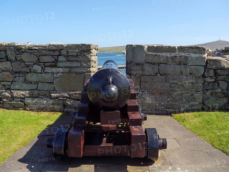 An old cannon pointing to the blue waters of the coast in front of a historic wall and standing on a patch of grass, Lerwick, Shetlands, Scotland, United Kingdom, Europe