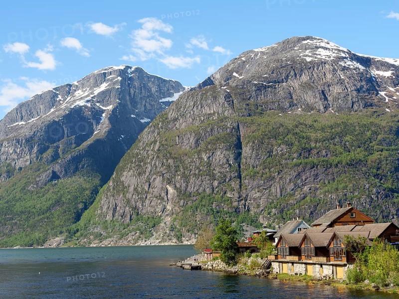 Wooden houses on the lakeshore in front of large, snow-covered mountains under a clear sky, Eidfjord NOR 3