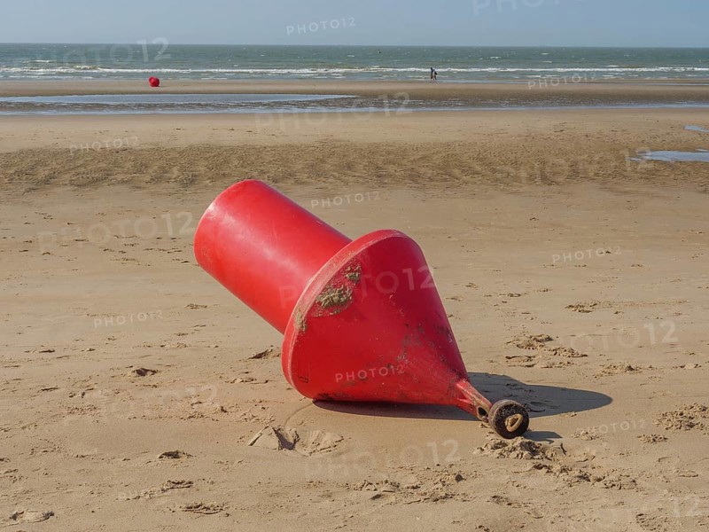 A large red buoy lies on the sand on the beach with the sea in the background, de haan, belgium