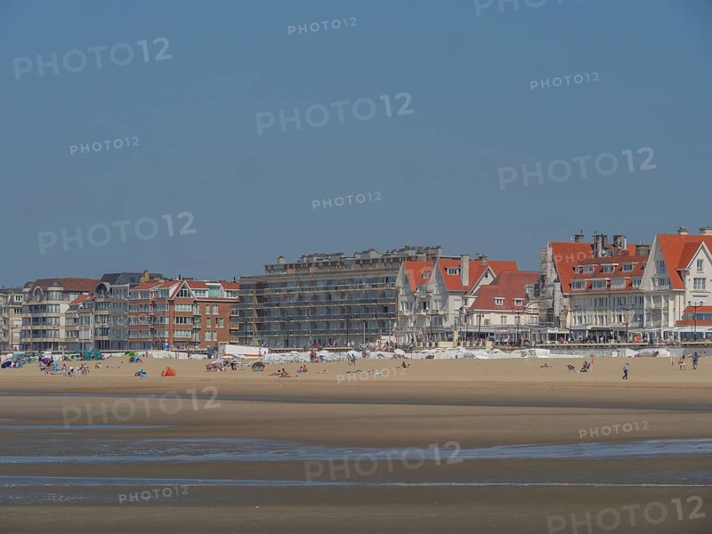Row of buildings along a busy sandy beach with holidaymakers and calm sea in the background under a clear sky, de haan, belgium