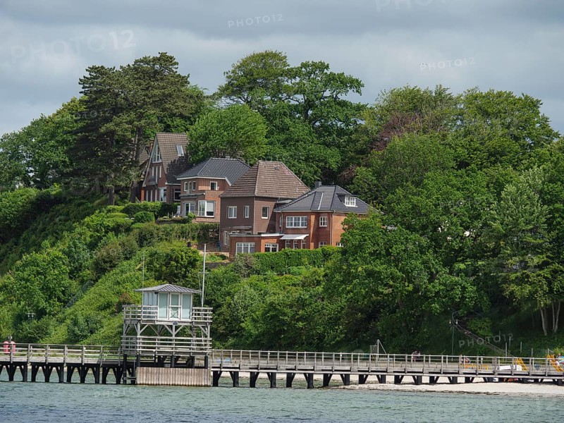 Houses on a green hill, surrounded by trees, with a footbridge leading into the water, under a partly cloudy sky, Glücksburg, Schleswig-Holstein, Germany, Europe
