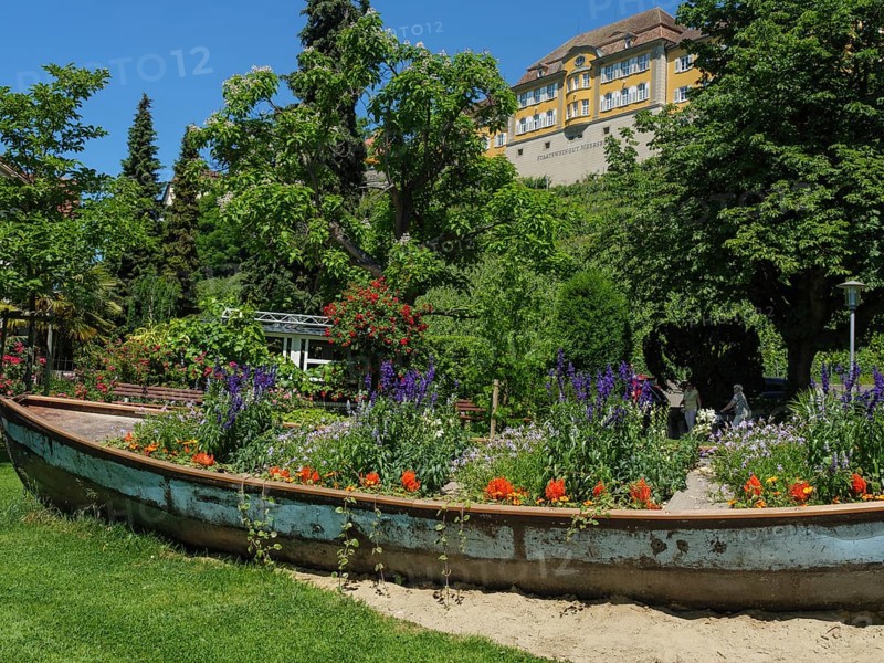 An old boat serving as a flower box in a green garden with a large yellow building in the background, Meersburg, Lake Constance, Germany, Europe