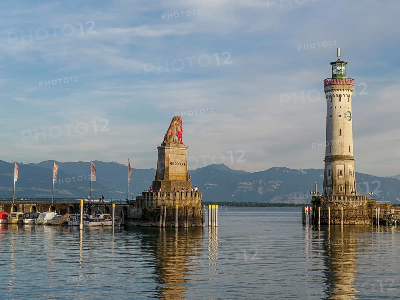 View of a harbour with a lion sculpture and a lighthouse against a mountain backdrop in the morning sun, Meersburg, Lake Constance, Germany, Europe