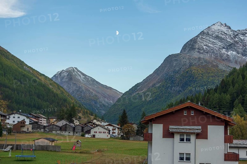 A mountain village with scattered houses in a valley between high mountains under a clear sky with a crescent moon, Saas Fee, Switzerland, Europe