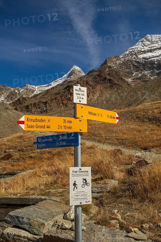 A signpost on a hiking trail in the mountains under a clear blue sky, surrounded by dry landscape elements, Saas Fee, Switzerland, Europe