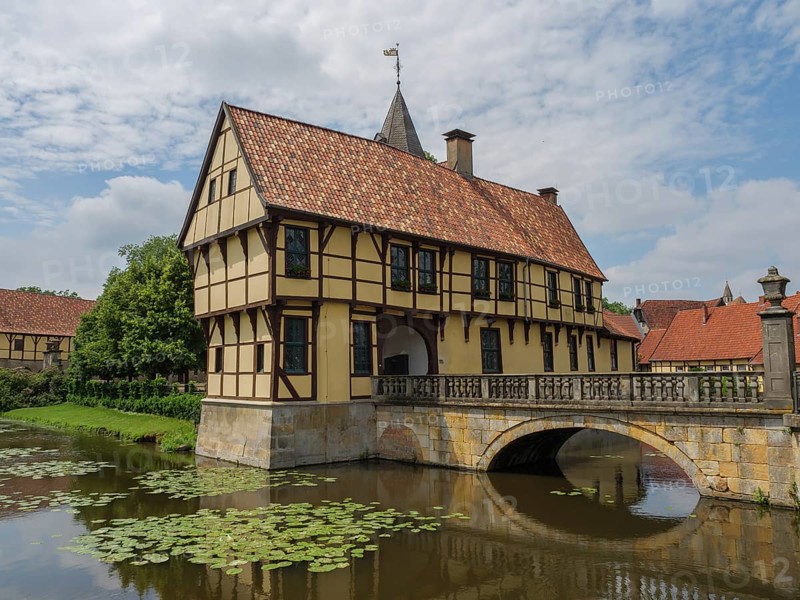 A half-timbered house with red roof tiles standing over a river with water lilies on a stone bridge under a cloudy sky, Steinfurt, North Rhine-Westphalia, Germany, Europe