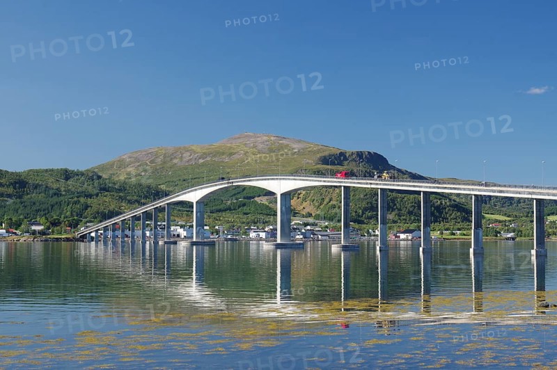 A curved bridge spans calm waters with reflective mountains and green landscapes under a clear sky, Sortland, Langoya, Vesteralen, Norway, Europe