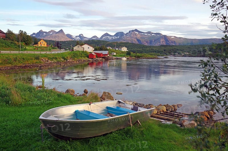 An abandoned boat lies on a grassy shore with mountains and houses in the background, Saltstraumen, Nordland, Norway, Europe