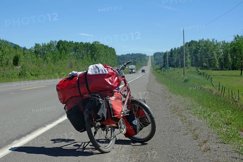 A bicycle loaded with panniers stands on the side of the road in a green, rural landscape under a blue sky, long endless degrees, bicycle tour, adventure holiday, British Columbia, Canada, North America