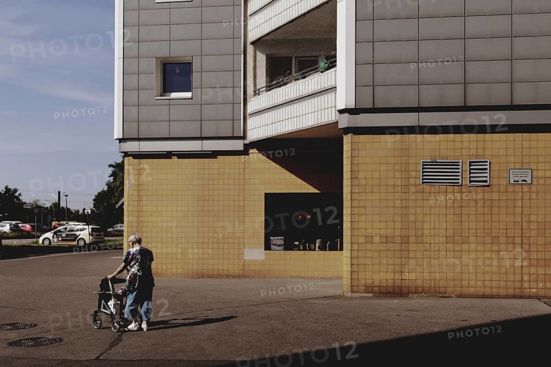 An old woman with a walking frame stands out in front of a block of flats in Marzahn, taken in Berlin, 07/08/2024
