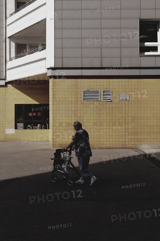 An old woman with a walking frame stands out in front of a block of flats in Marzahn, taken in Berlin, 07/08/2024