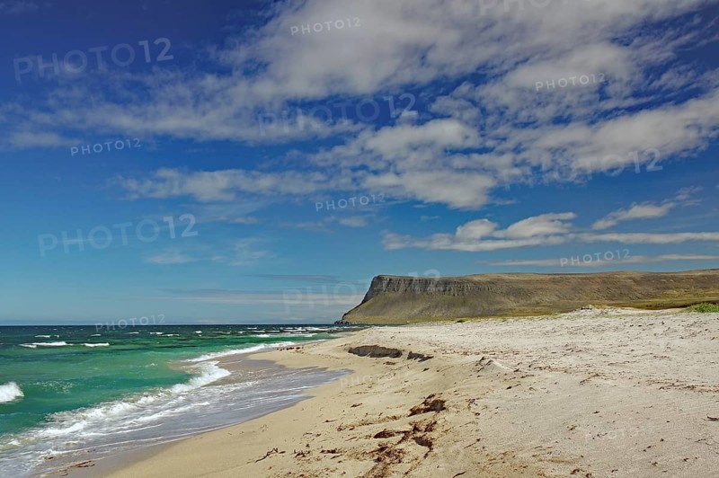 A sunny beach with deep blue water and waves, surrounded by cliffs and bright skies, Latrabjarg, Vestfirdir Peninsula, Iceland, Europe