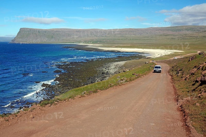 A car drives along a coastal road passing a rugged coastline with blue water, Latrabjarg, Vestfirdir Peninsula, Iceland, Europe