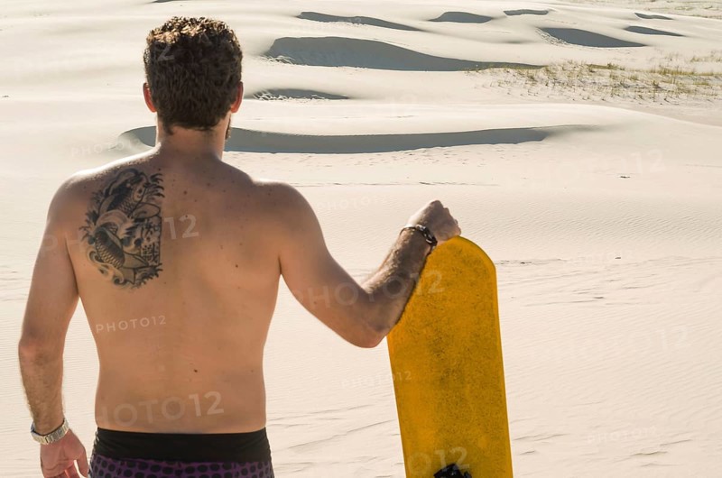 Young man on his back looking at the sand dunes, preparing to practice sandboarding