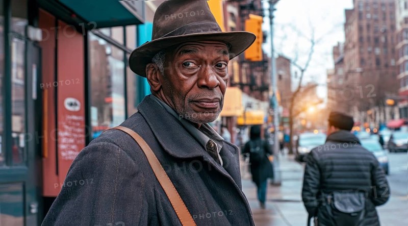 Senior black man wearing a hat and a coat standing on a sidewalk of a black neighborhood community, AI generated