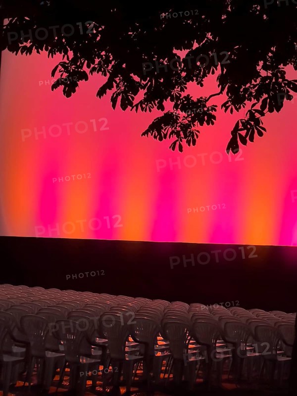 Empty rows of seats in the open-air cinema in front of a brightly lit stage, illuminated in red and orange, under a tree