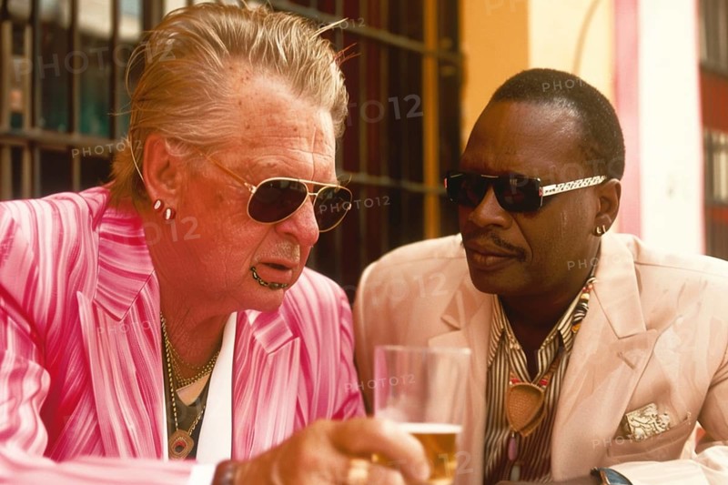 Two elderly men in striped suits with sunglasses enjoy a relaxed conversation over beer at an outdoor cafe table on a sunny day, AI generated