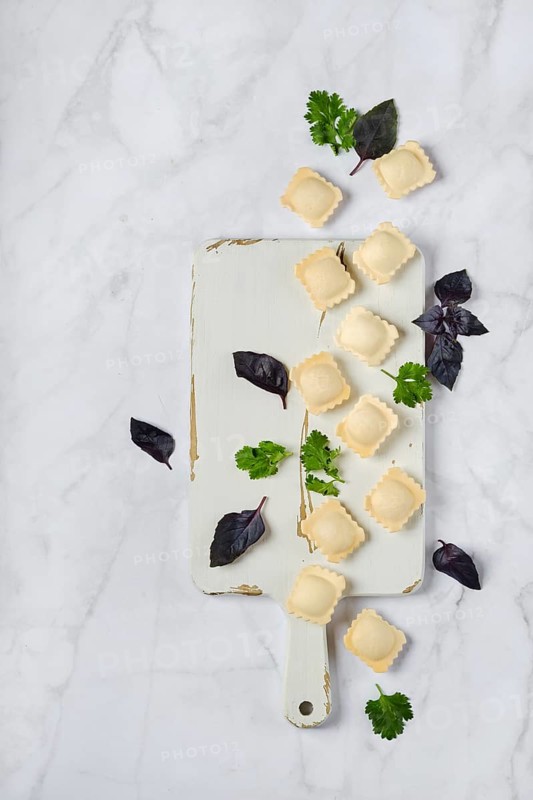 Overhead view of raw uncooked ravioli on a chopping board with scattered leaves of basil and cilantro