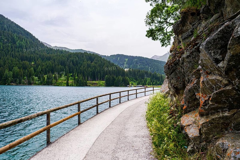 A winding path along the lake with a wooden railing and rocky side, surrounded by wooded mountains, Davos, Switzerland, Europe