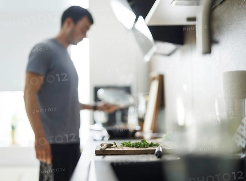 Cooking theme. A chopping board with parsley, in the background a man stands at the cooker and lifts the lid of a pan. Berlin, 01.08.2024